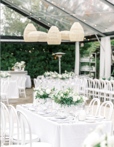 Elegant outdoor wedding setup with white chairs and tables under a clear tent. Tables decorated with white floral centerpieces; wicker pendant lights hang above.